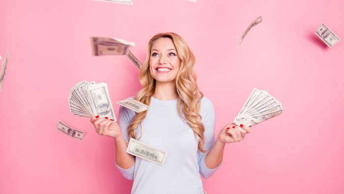 Portrait of pretty charming positive cute successful lucky cheerful girl standing under shower from money having a lot of money in hands isolated on pink background