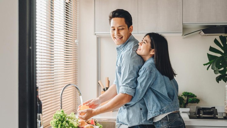 Asian couple lovers hug in the kitchen while cooking at home. A woman hugs a man who is washing vegetables from the back.