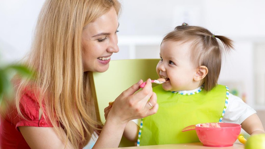 Happy mother spoon feeding her baby child in nursery