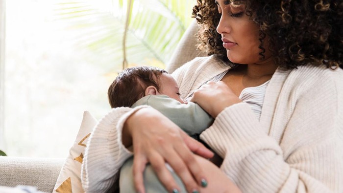 Sitting on a chair in the living room, the new mom watches as her baby boy breastfeeds.