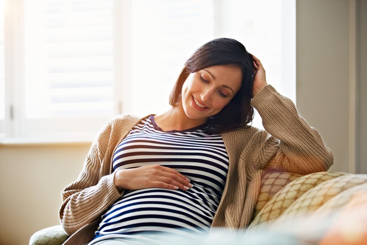 kehamilan Shot of a smiling pregnant woman sitting on her sofa holding her stomach