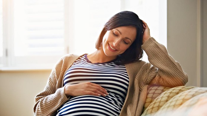 Shot of a smiling pregnant woman sitting on her sofa holding her stomach