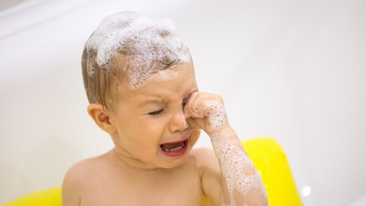 Cute toddler in tears crying taking a bath in bathtub, scared kid afraid of water in bathroom touching irritated by shampoo foam eyes, sad emotional baby screaming weeping in tantrum while bathing