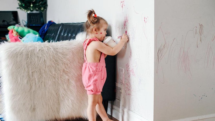Funny cute baby girl drawing with marker on wall at home. Toddler girl child with milk bottle playing at home. Authentic candid childhood lifestyle moment. Young artist painting on wall in living room