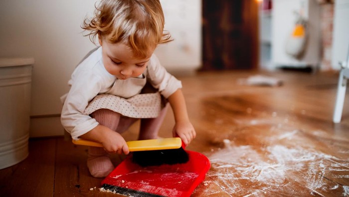 A small toddler girl with brush and dustpan sweeping messy floor in the kitchen at home.