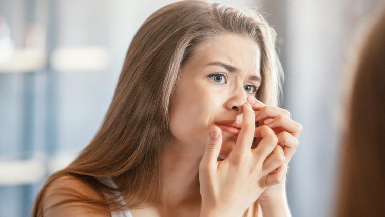 Displeased young woman squeezing acne on her nose while looking in mirror at home