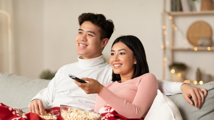 Asian Spouses Enjoying Christmas Eve Watching TV And Xmas Movies Sitting On Couch In Living Room At Home. Asian Family Watches Television Indoors. New Year And X-Mas Celebration Concept