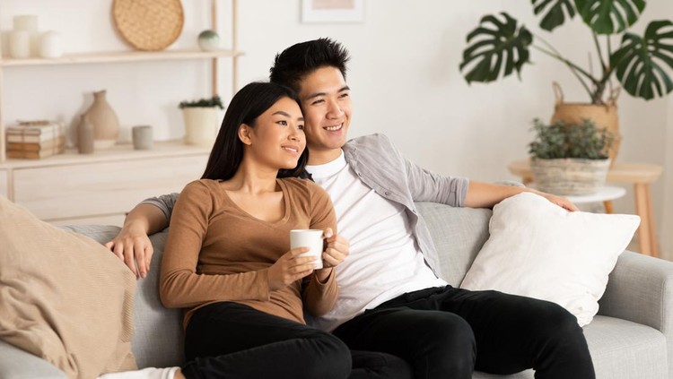 Asian Spouses Enjoying Christmas Eve Watching TV And Xmas Movies Sitting On Couch In Living Room At Home&period; Asian Family Watches Television Indoors&period; New Year And X-Mas Celebration Concept