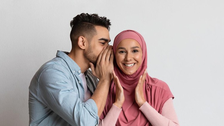 Pasangan suami istri Gossips Concept. Cheerful arab guy whispering something into his muslim girlfriend's ear, millennial man sharing secret with islamic woman in hijab, having fun over grey studio background, free space