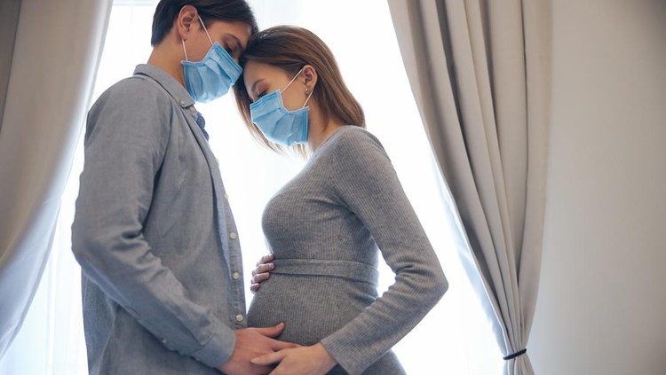 Man and woman in medical masks. Calm young pregnant lady leaning towards her husband and touching her belly