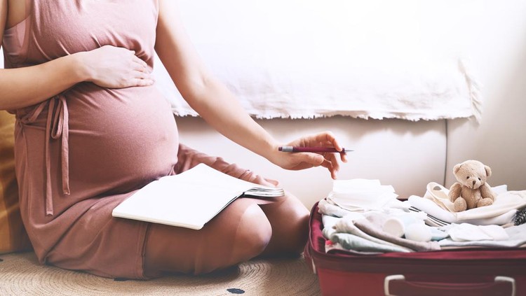 Pregnant woman packing bag for maternity hospital, making notes, checking list in diary. Expectant mother with suitcase of baby clothes and necessities preparing for newborn birth during pregnancy.