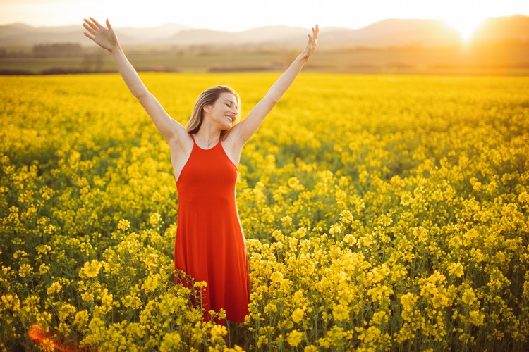 warna keberuntungan zodiak Beautiful young woman wearing bright red dress in yellow canola plant field. The field is spacious and the sun is showing in the background above the nearby hill. She is smiling and raising arms to the sky. Enjoying and celebrating freedom, happiness and positive thoughts and life.