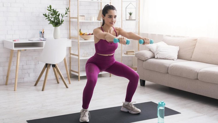 At home sports. Latin girl doing squats with dumbbells in living room