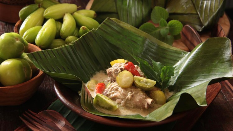 Garang Asem Ayam, the traditional Javanese tangy chicken curry. Each portion is placed in a banana leaf pouch then steamed. One portion is served and plated on an earthenware plate. Some fresh green tomatoes, bilimbi fruits, and unopened pouches are arranged on the table.