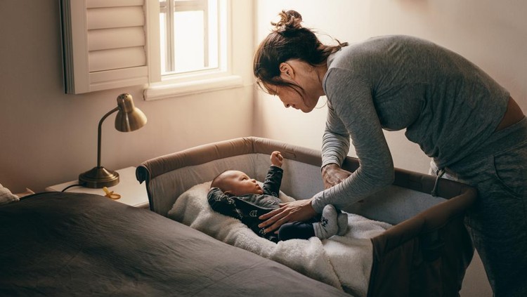 Mother putting her baby to sleep on a bedside baby crib. Woman bending forward over a crib to check her sleeping baby.