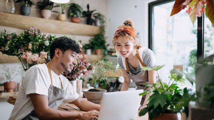 Smiling young Asian couple, the owners of small business flower shop, discussing over laptop on counter against flowers and plants. Start-up business, business partnership and teamwork. Working together for successful business