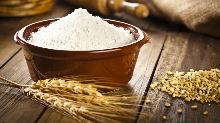Bowl filled with wheat flour on a rustic wooden table. Bowl is on left side and on right side there is a heap of whole wheat seeds.  In front of the bowl, there are three wheat ears. Shot with a DSLR Canon EOS 5D Mark II