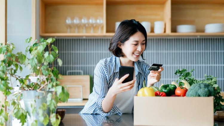 Beautiful smiling young Asian woman grocery shopping online with mobile app device on smartphone and making online payment with her credit card, with a box of colourful and fresh organic groceries on the kitchen counter at home