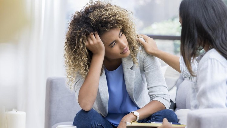 1026611056 A young woman sits on a couch with her unrecognizable therapist. She puts her head in her hand as she looks out the window with a sad expression. Her therapist puts a hand on her shoulder.
