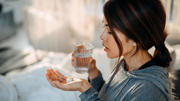 Young Asian woman sitting on bed and feeling sick, taking medicines in hand with a glass of water
