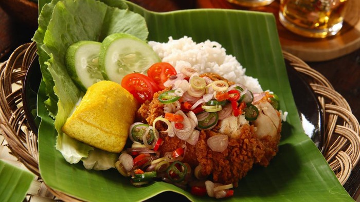 Ayam Geprek Sambal Matah, the popular fusion street food dish of southern fried chicken topped with Sambal Matah (salsa of chili pepper, lemon grass and shallot) from Bali. The chicken and condiment are served with steamed rice, yellow tofu, and fresh vegetables. The dish is plated on a ceramic plate lined with banana leaf; which then placed on a woven stick plate.