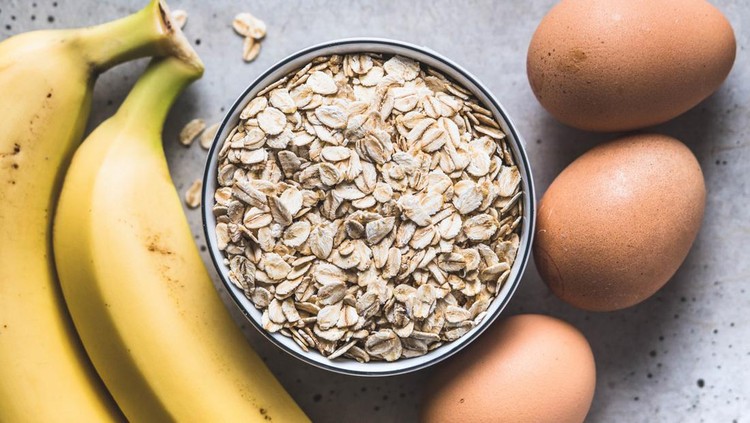 A bowl with oatmeal next to bananas and eggs on a concrete table. Healthy pancakes ingredients.