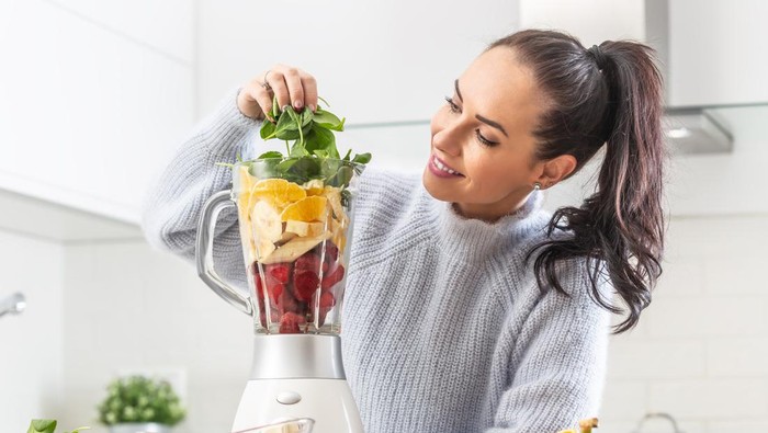 Good looking girl piles colorful mix of fruit and green leaves into a blender at home in the kitchen.