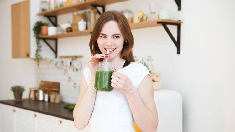Smiling young woman drinking green smoothie juice in kitchen. Healthy Lifestyle.