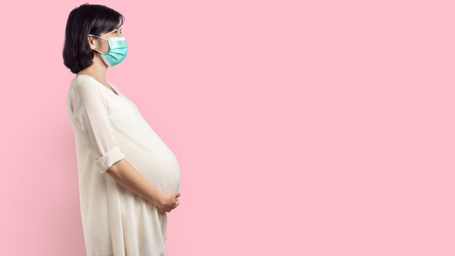 Happy Smiling Asian Pregnant woman wearing protective face mask standing over pink isolated background. Expectant female holding her belly with copy space.