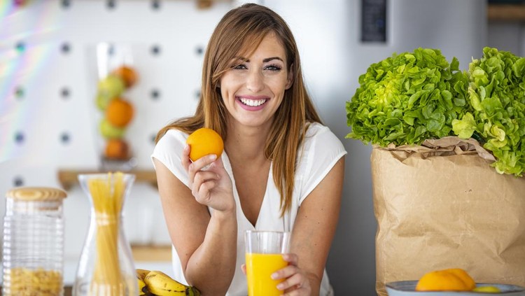 Young joyful woman drinking orange juice and standing near a kitchen table. Close up of a woman drinking juice in her kitchen. Fit smiling young woman preparing healthy fruit juice