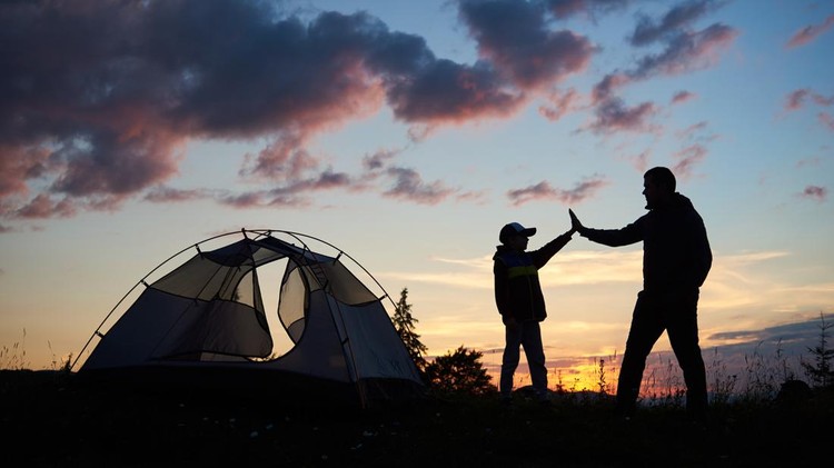Silhouette of man and child hikers give each other a high five near the tent at dawn against the backdrop of the mountains and the sun rising above them. Concept of healthy lifestyle.