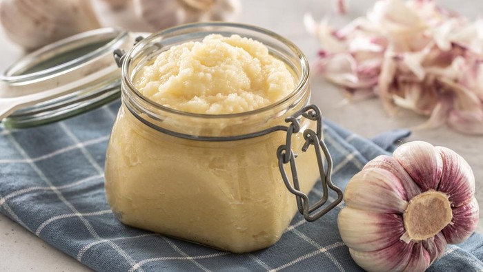 Aromatic garlic paste in a glass jar laid on rustic kitchen cloth with bulbs and peeled cloves.