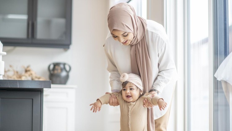 Ibu Muslimah A Muslim mother is encouraging her baby girl to learn how to walk. She is holding her daughter by her arms and attempting to help her stand on her feet.