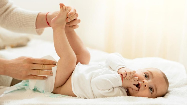 ilustrasi ganti popok Baby boy lying on the bed with hands in the mouth while getting his diaper changed.