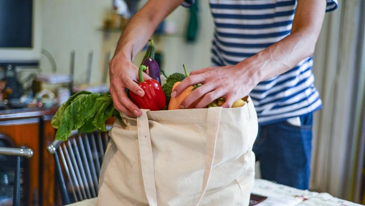 Asian man unpacking groceries at kitchen island. He is removing fruits and vegetables from reusable bags.