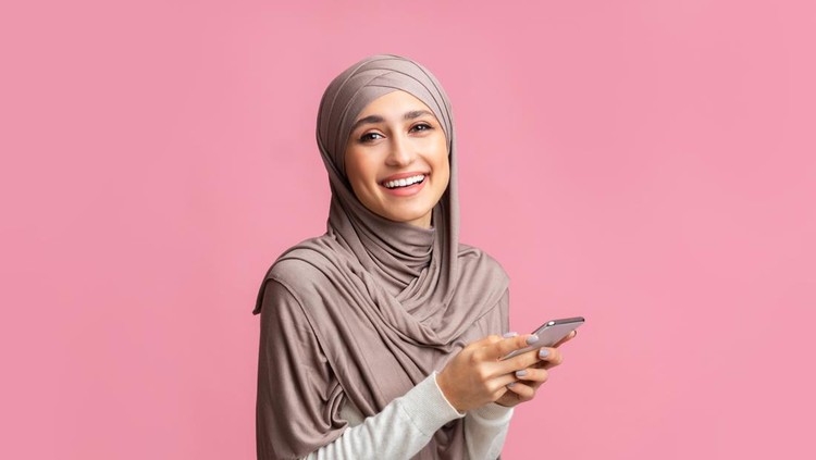 Ilustrasi Bisnis Ramadan 2022 Cool application. Happy muslim girl in hijab using smartphone, texting or browsing internet and looking at camera, pink background with free space