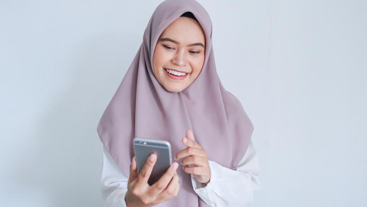 Young Asian Islam woman wearing headscarf is smile and happy in what she see on the smartphone. Indonesian woman on gray background.