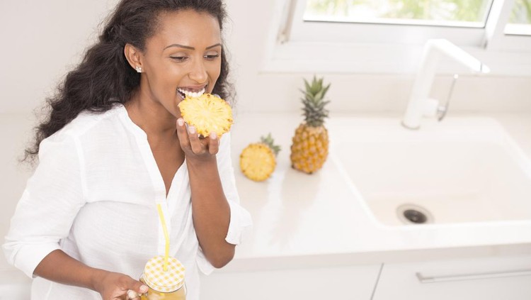 Happy woman eating pineapple and drinking freshly squeezed pineapple juice. Attractive Ethiopian woman wearing a white shirt, standing, leaning on the kitchen countertop.