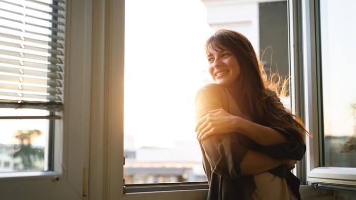 Beautiful young woman looking through the window while enjoying fresh air