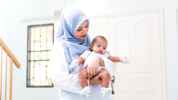 A young girl holds her baby brother in her arms while helping out her mother in the kitchen.  She is dressed casually and is wearing a Hijab.  There are dishes out on the counter as she works about in the kitchen.