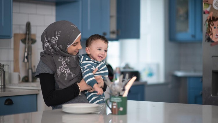 A young girl holds her baby brother in her arms while helping out her mother in the kitchen.  She is dressed casually and is wearing a Hijab.  There are dishes out on the counter as she works about in the kitchen.