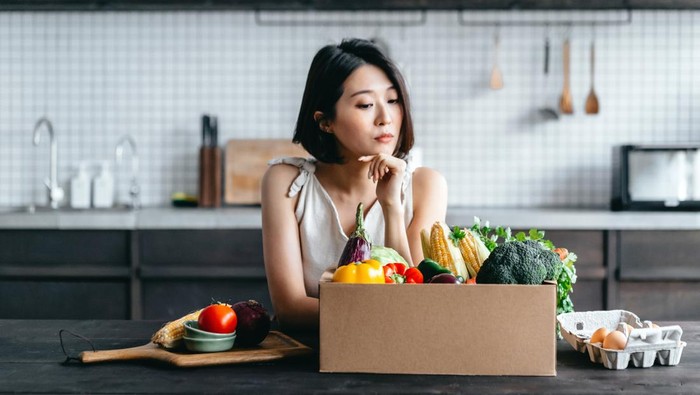 Young Asian woman received a box full of colourful and fresh organic groceries ordered online by home doorstep delivery. She is planning to cook a healthy meal in the kitchen at home. Healthy eating lifestyle