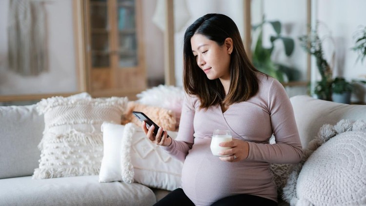 Beautiful young Asian pregnant woman relaxing on sofa in the living room at cozy home. Drinking a glass of fresh milk and using smartphone. Wellbeing, healthy eating lifestyle during pregnancy