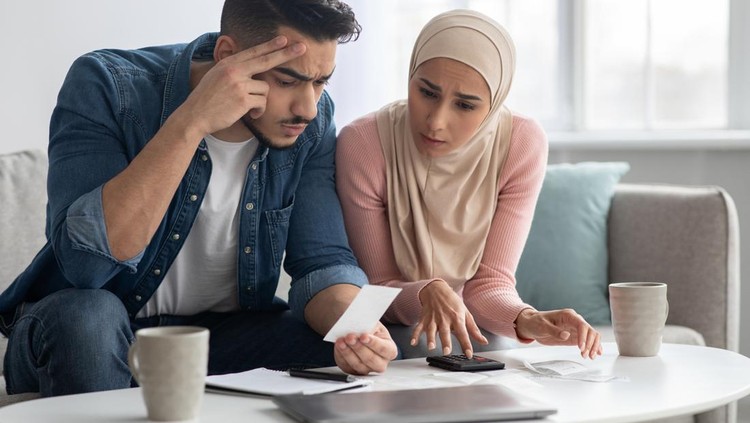 Confusded lady in hijab and her upset husband with bills, sitting together on couch in living room, middle-eastern young family counting their spendings, suffering from financial difficulties