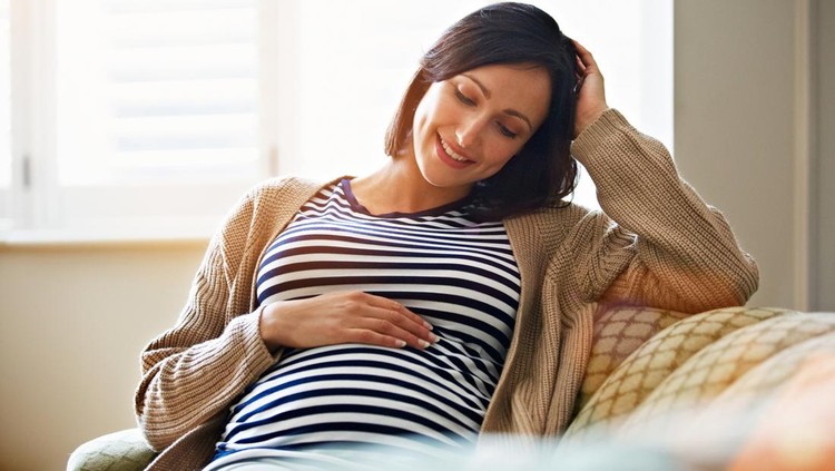 Shot of a smiling pregnant woman sitting on her sofa holding her stomach