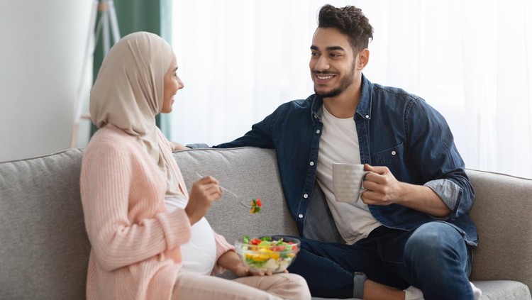 Pregnant Islamic Woman Eating Fresh Vegetable Salad While Resting On Couch With Husband, Happy Middle Eastern Family Awaiting For Baby Sitting Together On Comfortable Sofa In Living Room, Free Space