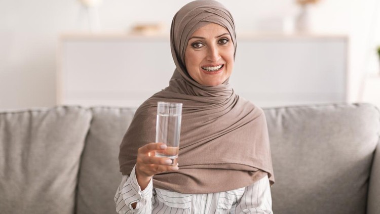 Happy Middle-Aged Muslim Woman In Hijab Posing Holding Glass Of Water Drinking Healthy Liquid For Hydration Sitting On Couch At Home, Smiling To Camera. Stay Hydrated Concept