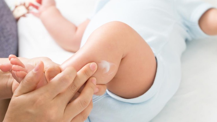 ruam bayi Young adult mother hand applying white medical ointment on toddler leg. Red rash on skin from diaper. Care about baby body. Closeup. Front view. Isolated on light gray background.