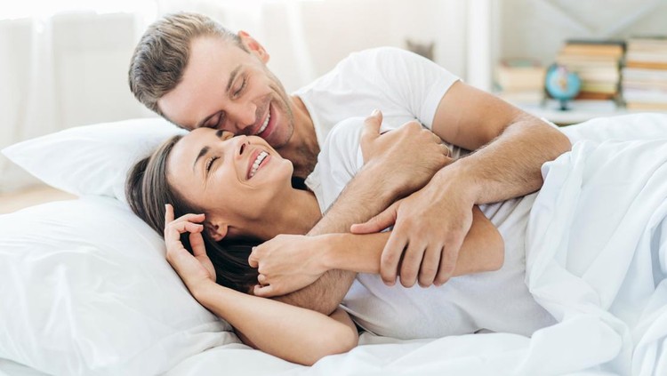Close up of young couple lying in bed together at the sunny morning. Man in woman in love smiling to each other and giving hugs.