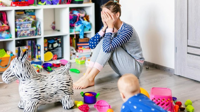 Tired of everyday household mother sitting on floor with hands on face. Kid playing in messy room. Scaterred toys and disorder.